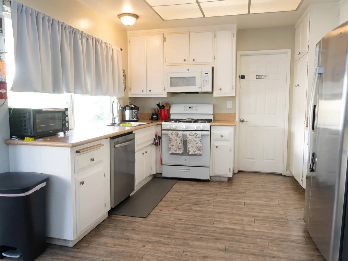 Spacious accessible kitchen with white cabinetry, wood flooring, and stainless steel appliances in residential care home